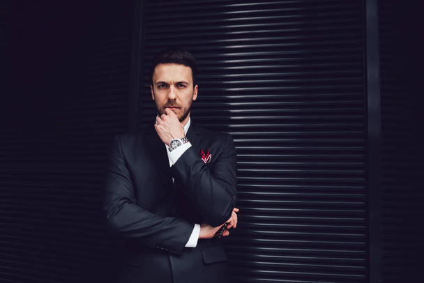 Serious pensive adult male entrepreneur in business suit looking at camera and touching face with hand while holding smartphone and standing at striped black wall  - Fotografie, Obrázek