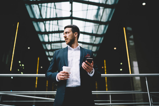 From below of bearded male manager in formal suit with coffee to go and mobile phone in hands looking away at modern office building  - Фото, изображение