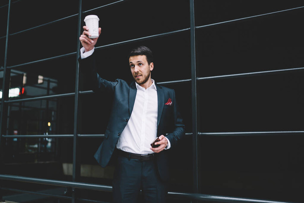 Confident handsome man in formal suit with smartphone in hand taking break drinking cup of coffee greeting coworker in urban street - Fotoğraf, Görsel