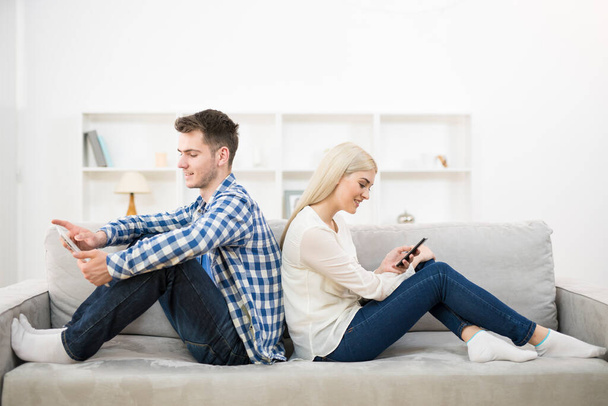 電話とタブレットを持った幸せな男と女がソファの上に座る - 写真・画像