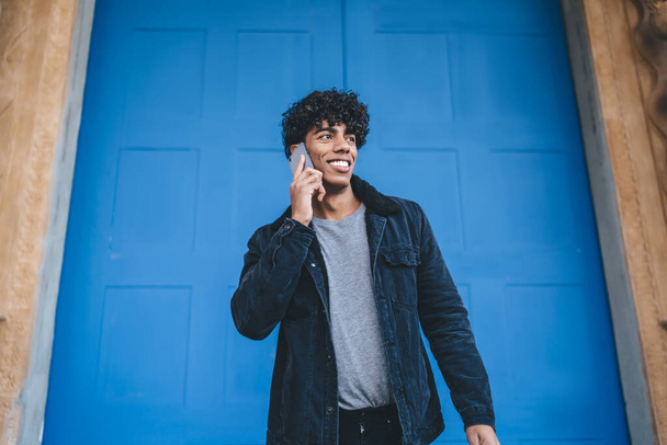 Smiling modern young curly ethnic man in black jacket and jeans having phone call while standing in front of urban blue door on street looking away  - Φωτογραφία, εικόνα