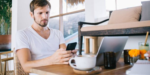 Focused confident man in white shirt resting on terrace with cup of coffee messaging on smartphone and working on laptop - Фото, зображення