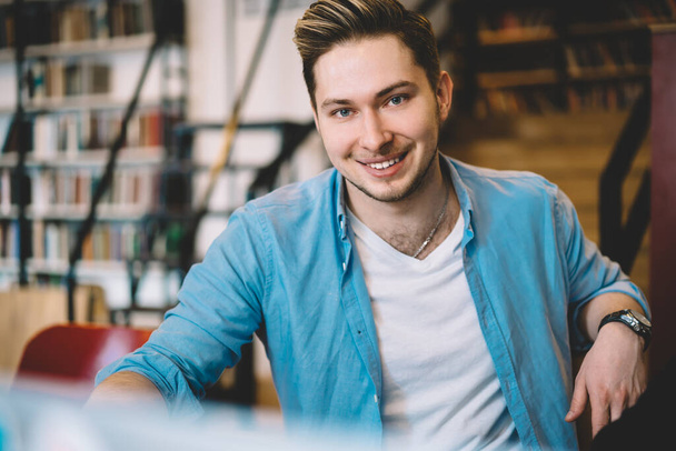 Portrait of cheerful handsome smiling male in casual clothes with watch spending time sitting at table in library looking at camera - Photo, image