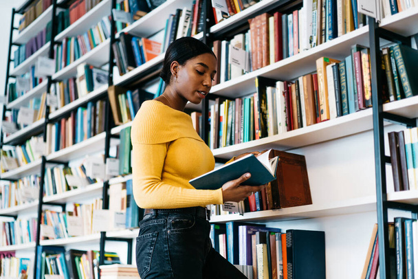 Focused African American lady in casual clothes reading textbook while standing near bookcase and studying in library of contemporary university - Zdjęcie, obraz