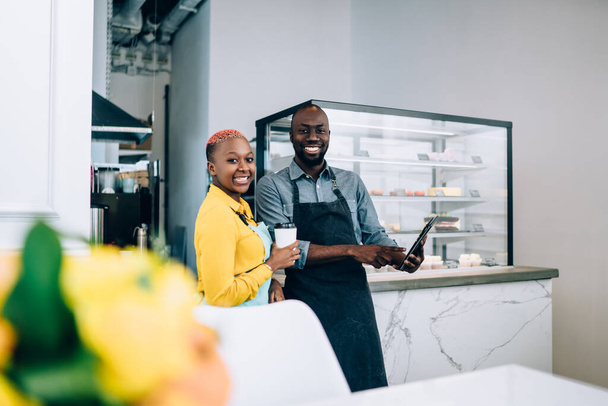 Cheerful smiling African American coworkers dressed in aprons standing with cup of coffee and tablet in cafe with counter with cakes in background - Zdjęcie, obraz
