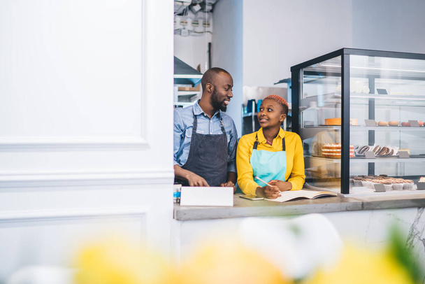 Young stylish African American woman in yellow shirt and blue aprons looking at black colleague and smiling while writing information on notebook behind counter of pastry shop - Фото, зображення