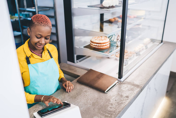 From above positive smiling African American young woman with short hairstyle in apron using tablet working standing at counter in pastry shop - Foto, Imagen
