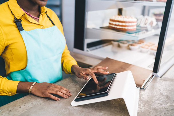 From above of crop African American female in apron doing work touching screen of tablet standing at counter in pastry shop - Fotografie, Obrázek
