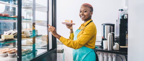 Side view of young smiling African American female worker of pastry shop pointing at  cake display fridge and holding plate with piece of cake while inviting for trying delicious sweets - Photo, Image