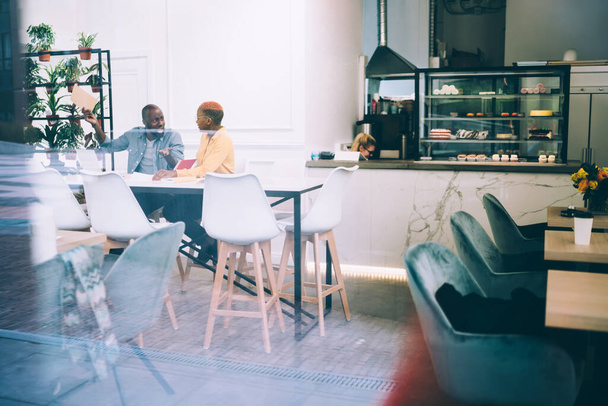 Focused African American coworkers in casual clothes working with documents sitting at table in coffee shop with counter with cakes in background - Фото, изображение