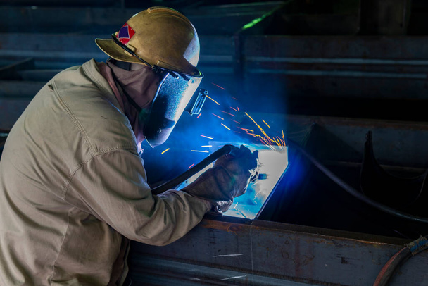 Ο συγκολλητής συγκολλεί μια δομή χάλυβα εργασία με τη διαδικασία Flux Cored Arc Welding (FCAW) και ντυμένος κατάλληλα με τον εξοπλισμό ατομικής προστασίας (PPE) για την ασφάλεια, στο βιομηχανικό εργοστάσιο. - Φωτογραφία, εικόνα