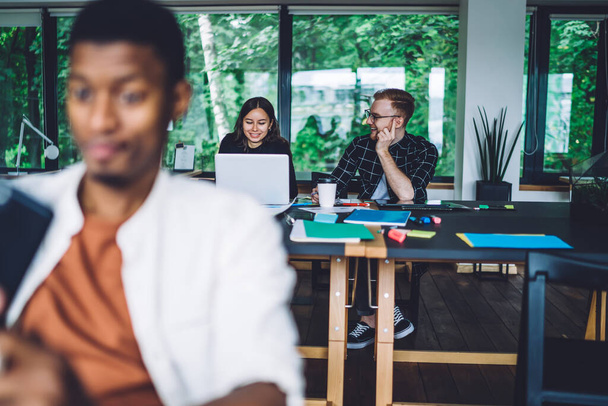 Glückliche smarte multiethnische Mitarbeiter in lässiger Kleidung surfen auf Laptop und Smartphone am Tisch mit einer Tasse Kaffee Dokumente Papiere im Büro - Foto, Bild