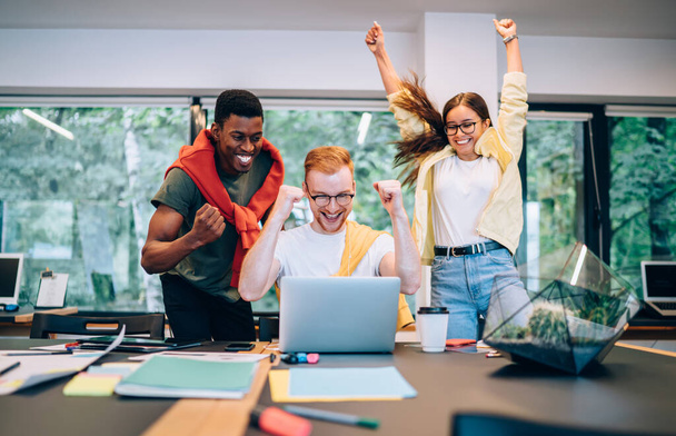 Group of excited diverse young people clenching fists and celebrating successful project fulfillment while working in modern creative workspace together - 写真・画像