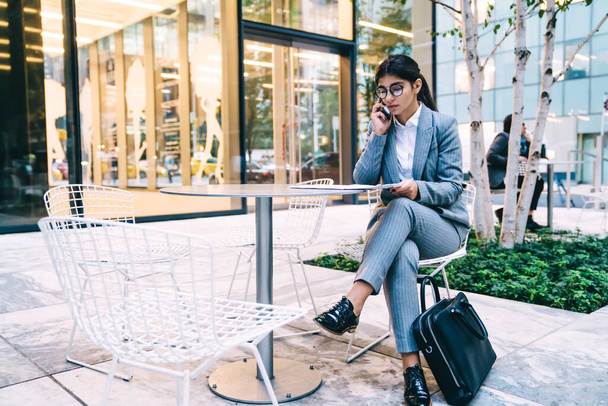 Giovane bella donna etnica indossa abito da ufficio e occhiali che lavorano con scartoffie in strada caffè parlando al telefono seduto a tavola su sfondo urbano - Foto, immagini