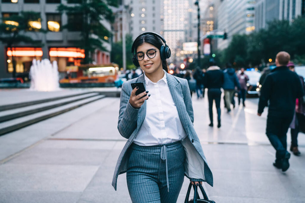 Joyful businesswoman in elegant formal clothing with round glasses using smartphone and listening to music while walking in downtown on blurred background - Φωτογραφία, εικόνα