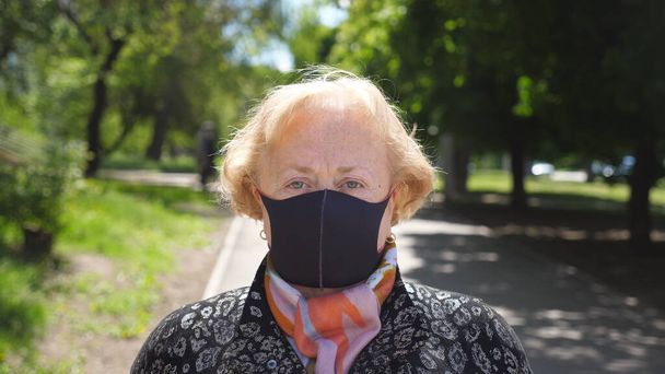 Portrait of old woman with black face mask stands at city street. Grandmother wears protective mask from virus COVID-19 outdoor. Concept of health and safety life from coronavirus pandemic. Close up - Photo, Image