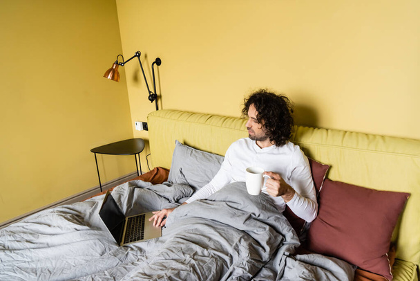 high angle view of curly freelancer using laptop with blank screen while holding cup of coffee in bed - Foto, Bild