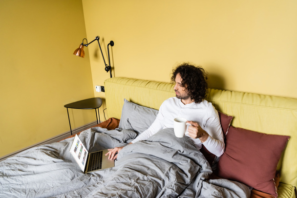 KYIV, UKRAINE - APRIL 25, 2020: high angle view of freelancer using laptop with Youtube website while holding cup of coffee in bed - Foto, Imagem