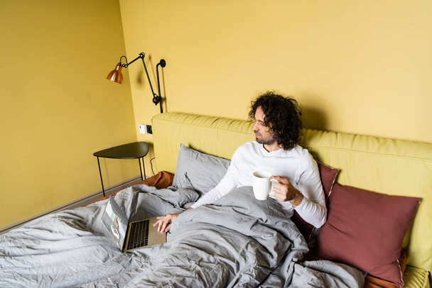 vysoký úhel pohledu na mladé freelancer pomocí notebooku s vstupenkami on-line webové stránky při držení šálek kávy v posteli - Fotografie, Obrázek