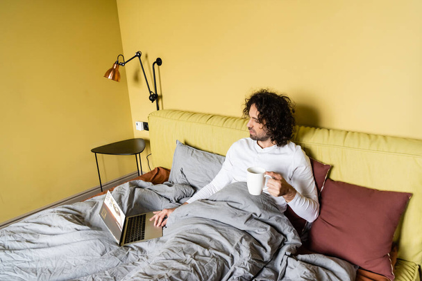 magas látószögű kilátás fiatal szabadúszó segítségével laptop jegyek online honlapon, miközben gazdaság csésze kávét az ágyban - Fotó, kép