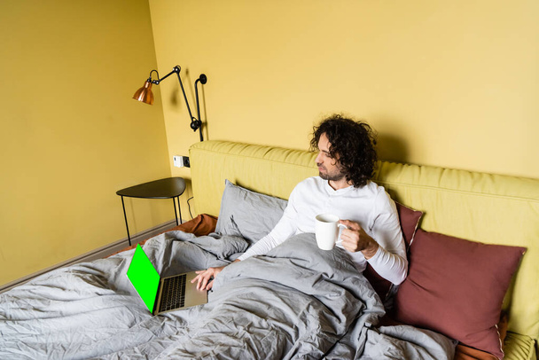ベッドでコーヒーを飲みながら緑の画面でノートパソコンを使う若いフリーランサーの高い角度からの眺め - 写真・画像