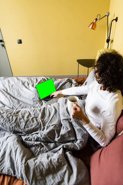 high angle view of curly freelancer using laptop with green screen while holding cup of coffee in bed - Foto, immagini