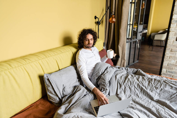 curly freelancer using laptop while holding cup of coffee in bed - Zdjęcie, obraz