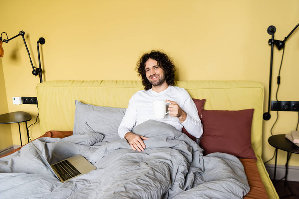 счастливый фрилансер за чашкой кофе, улыбаясь фотоаппарату в постели рядом с ноутбуком
 - Фото, изображение