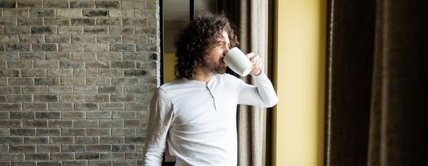 οριζόντια εικόνα του όμορφου άνδρα με πιτζάμες πίνοντας καφέ, ενώ κοιτάζοντας έξω από το παράθυρο το πρωί - Φωτογραφία, εικόνα
