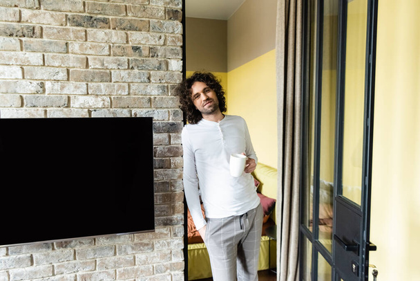 hombre guapo en pijama sosteniendo taza de café y mirando a la cámara cerca de pantalla LCD en blanco colgando en la pared de ladrillo
 - Foto, imagen