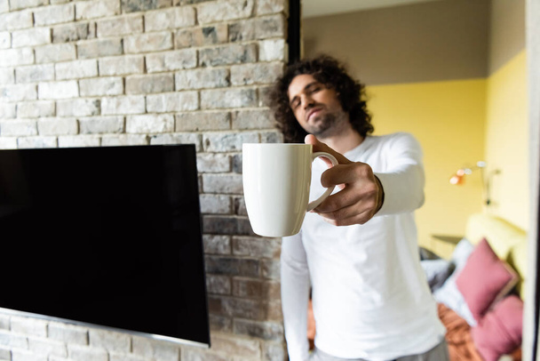 επιλεκτική εστίαση του όμορφου άνδρα κρατώντας φλιτζάνι καφέ σε τεντωμένο χέρι κοντά κενό οθόνη LCD κρέμεται στον τοίχο από τούβλα - Φωτογραφία, εικόνα