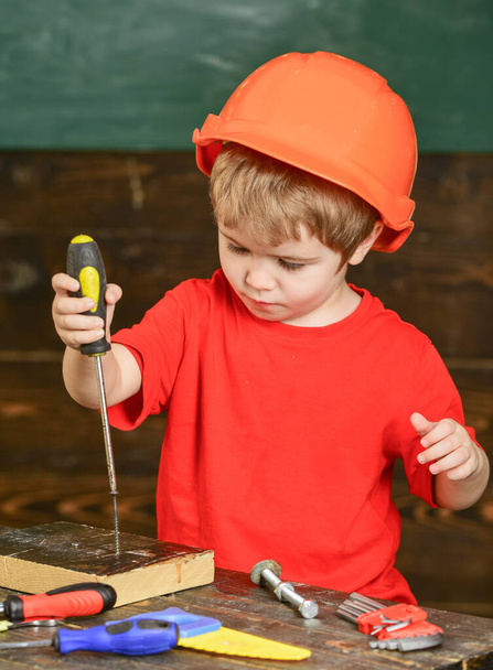 Kid boy holds screwdriver tool. Toddler on busy face plays with screwdriver at workshop. Handcrafting and workshop concept. Child in helmet cute playing as builder or repairer, or handcrafting. - Foto, imagen
