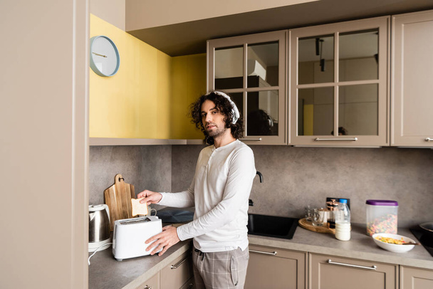 Kablosuz kulaklıklı kıvırcık adam mutfakta kameraya bakarken ekmek kızartma makinesine ekmek koyuyor. - Fotoğraf, Görsel