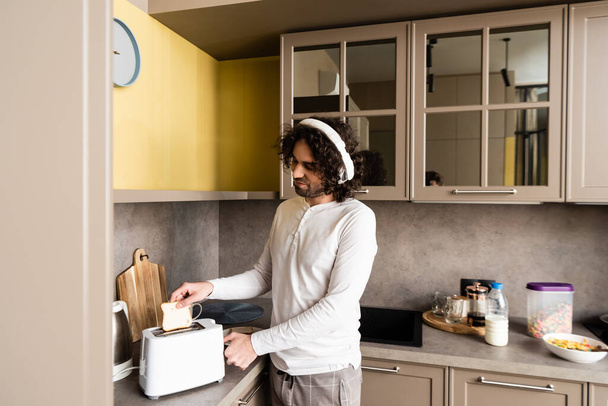 homem encaracolado em fones de ouvido sem fio colocando pão na torradeira enquanto prepara o café da manhã
 - Foto, Imagem