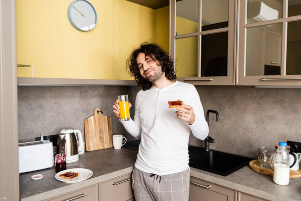 szczęśliwy człowiek w piżamie trzymając sok pomarańczowy i tosty z dżemem uśmiechając się do kamery - Zdjęcie, obraz