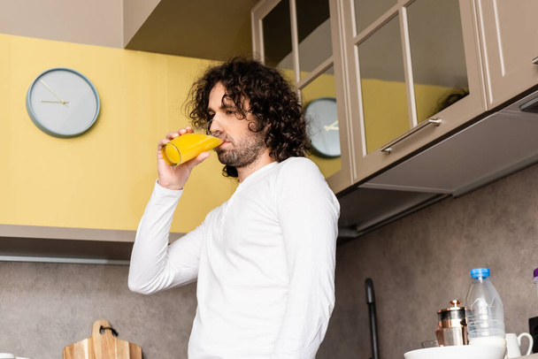 Düşük açılı, düşünceli bir adamın mutfakta dikilirken portakal suyu içmesi. - Fotoğraf, Görsel
