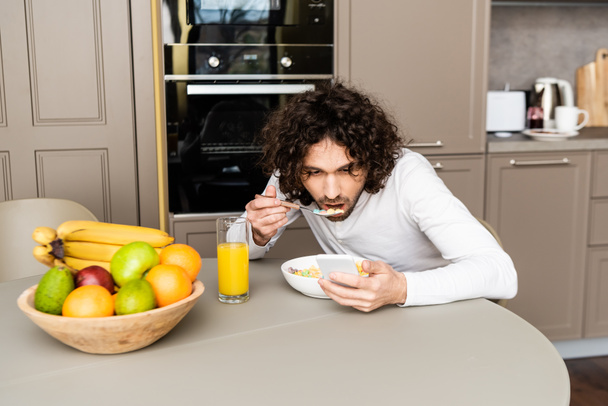 концентрований, кучерявий чоловік спілкується на смартфоні під час сніданку на кухні біля миски з фруктами
 - Фото, зображення