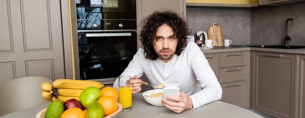 imagem horizontal de homem sério olhando para a câmera enquanto toma café da manhã e conversa no smartphone na cozinha
 - Foto, Imagem