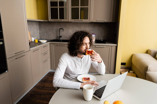 przystojny freelancer jedzenie tosty podczas korzystania z laptopa w pobliżu filiżanki kawy w kuchni - Zdjęcie, obraz