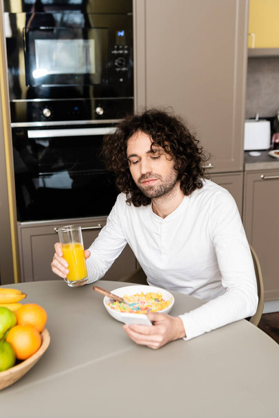 knappe, krullende man chatten op smartphone tijdens het ontbijt in de keuken - Foto, afbeelding