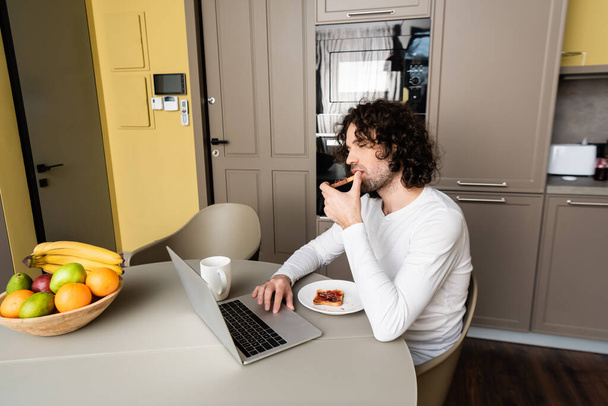thoughtful freelancer eating toast while looking at laptop near coffee cup and fresh fruits - Foto, Imagen