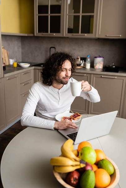 selective focus of freelancer drinking coffee near laptop, toasts and fresh fruits - Foto, Imagem
