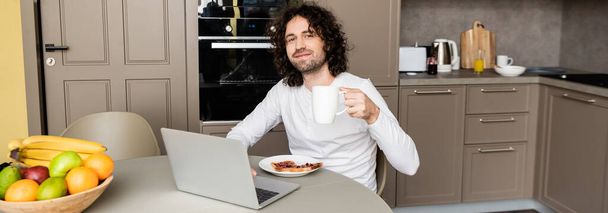 vízszintes kép mosolygós szabadúszó kezében kávéscsésze és nézi kamera közelében laptop, pirítós és friss gyümölcs - Fotó, kép