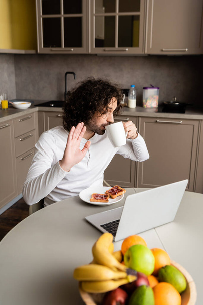 curly freelancer drinking coffee and waving hand during video chat on laptop in kitchen - Photo, Image