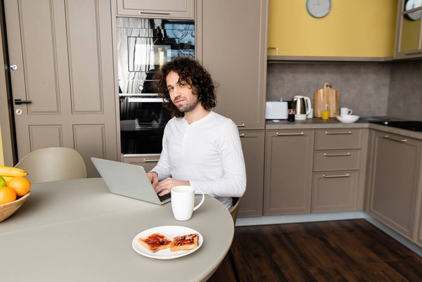 young blogger looking at camera while typing on laptop near plate with tasty toasts and coffee cup - Foto, afbeelding
