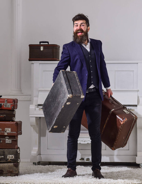 Man, traveller with beard and mustache with luggage, luxury white interior background. Macho elegant on smiling face stands near pile of vintage suitcase, holds suitcases. Baggage delivery concept. - Foto, immagini