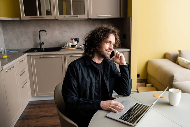 freelancer sorrindo falando no smartphone perto de laptop e xícara de café na cozinha
 - Foto, Imagem