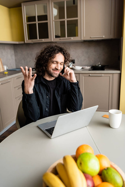 selective focus of smiling freelancer gesturing while talking on smartphone near fresh fruits and coffee cup - Fotoğraf, Görsel