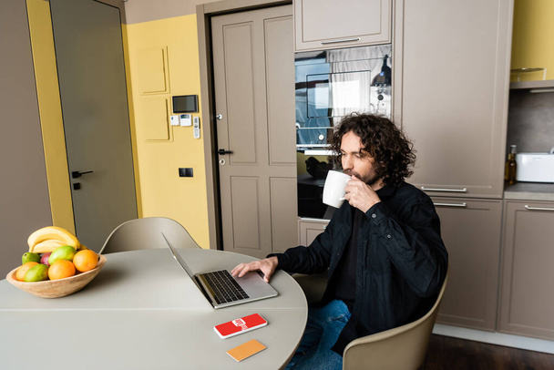 KYIV, UKRAINE - APRIL 25, 2020: handsome freelancer drinking coffee while using laptop near smartphone with Youtube app, credit card and fresh fruits - Foto, imagen