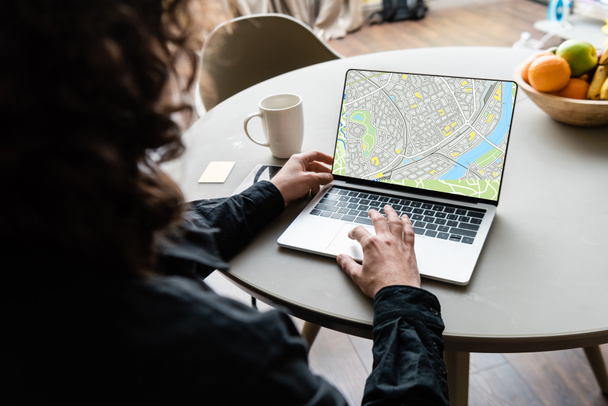 visão traseira do freelancer usando laptop com mapa na tela perto de xícara de café, notas pegajosas e frutas
 - Foto, Imagem